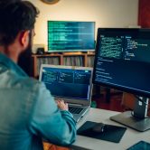 Rear view of a full-stack developer typing code on his laptop at Back view of a full-stack developer programming on a laptop at his home surrounded by two monitors. A senior developer is sitting at his home office and typing code on a laptop.