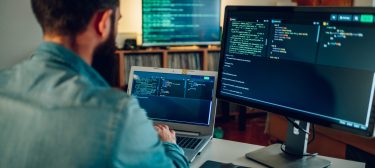 Back view of a full-stack developer programming on a laptop at his home surrounded by two monitors. A senior developer is sitting at his home office and typing code on a laptop.