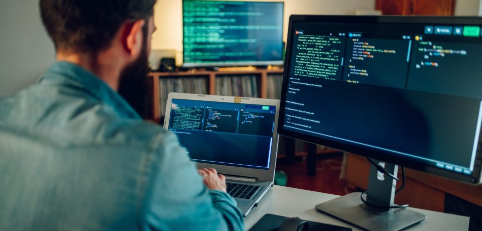 Rear view of a full-stack developer typing code on his laptop at Back view of a full-stack developer programming on a laptop at his home surrounded by two monitors. A senior developer is sitting at his home office and typing code on a laptop.