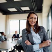 Young successful businesswoman at corporate office looking at camera. Confident businesswoman with arms crossed in the office looking at camera. Her colleagues are in the background.