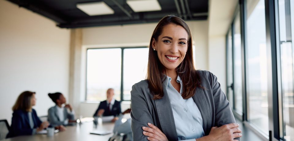 Young successful businesswoman at corporate office looking at camera. Confident businesswoman with arms crossed in the office looking at camera. Her colleagues are in the background.